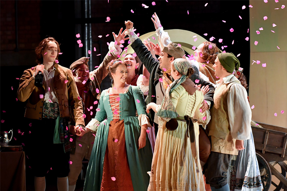 Students dressed up in historical costume, with two people being petals surrounding them, performing in an opera on stage.